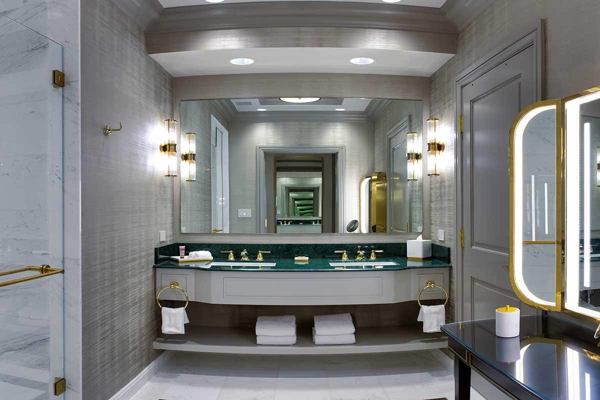 A hotel bathroom with a dual-sink vanity, a shower on the left, and a lighted mirror and table on the right.