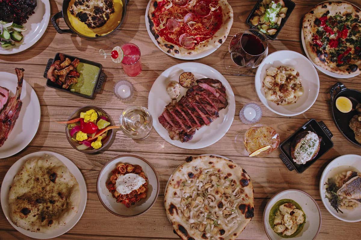 A variety of dishes on a table, including red meat, fish, and pizzas, and cocktails.