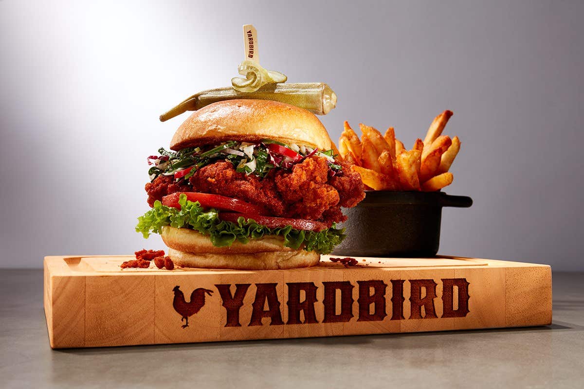 Fried chicken sandwich with lettuce, tomato, and pickle on a wooden board labeled "Yardbird," served with a side of fries.