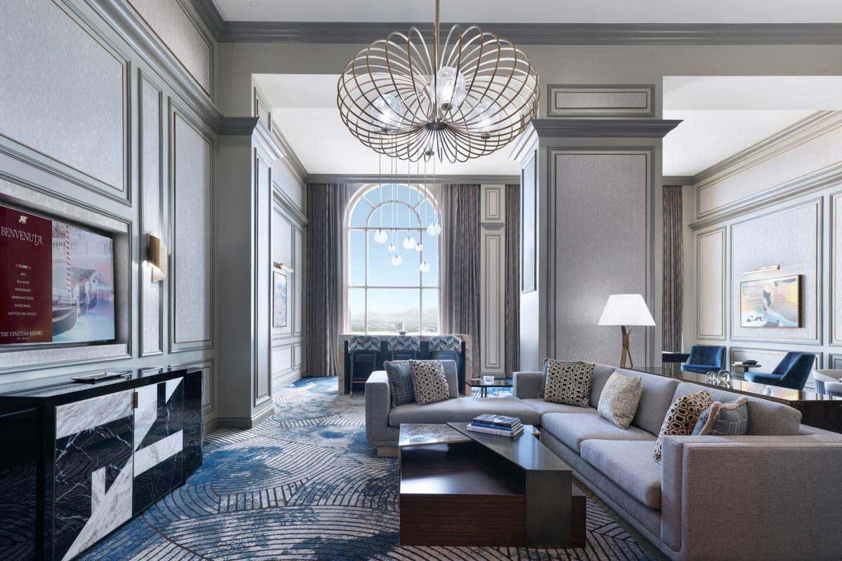 A hotel living room with a couch, coffee table, TV, a high-top table with bar chairs, chandeliers, armchairs, high ceilings, a tall arched window, and the edges of a pool table and dining table visible to the right.