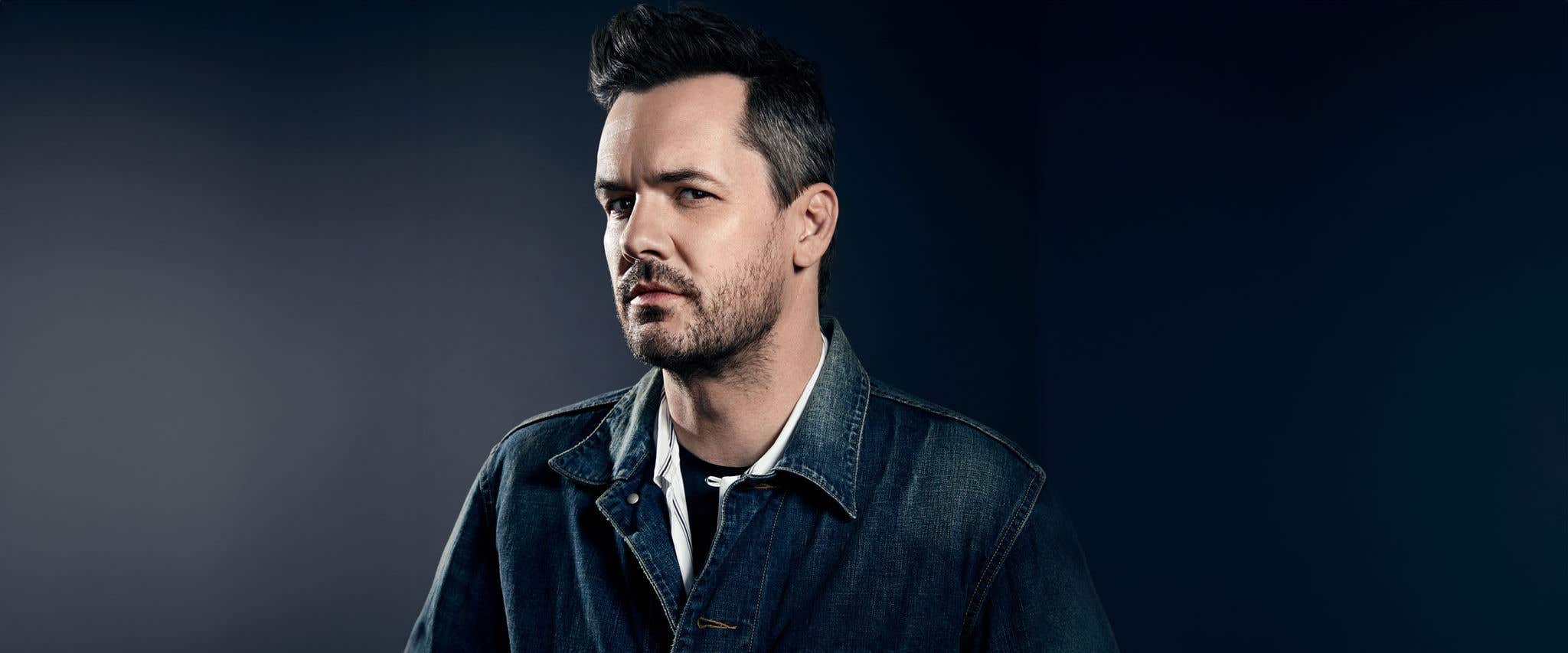 Close-up portrait of Jim Jefferies with short dark hair and light beard, wearing a denim jacket.