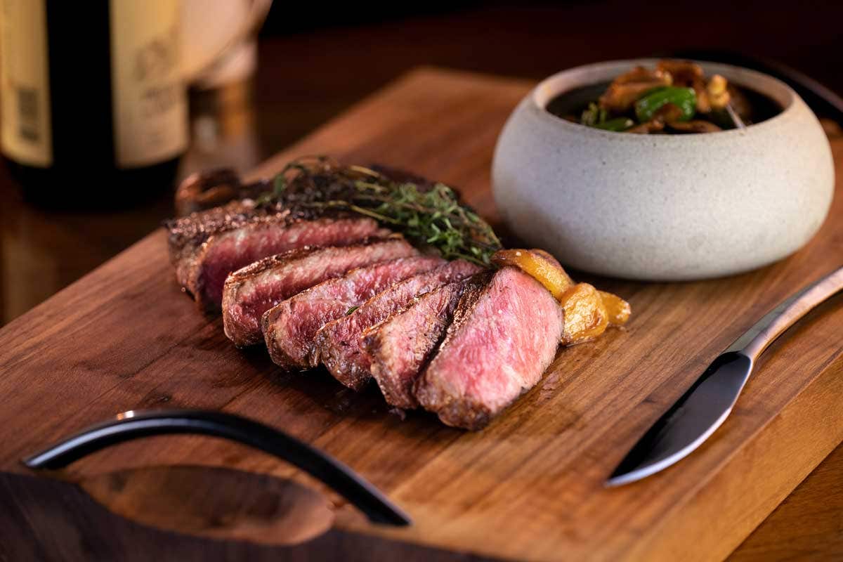 Medium-rare steak slices on a wooden board with herbs and a side dish in a gray bowl.