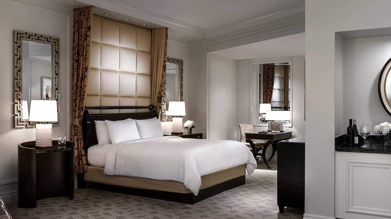 Hotel room with a king bed, white linens, two bedside tables with lamps, and a desk with a chair. Beige walls and carpet and decorative mirrors visible.