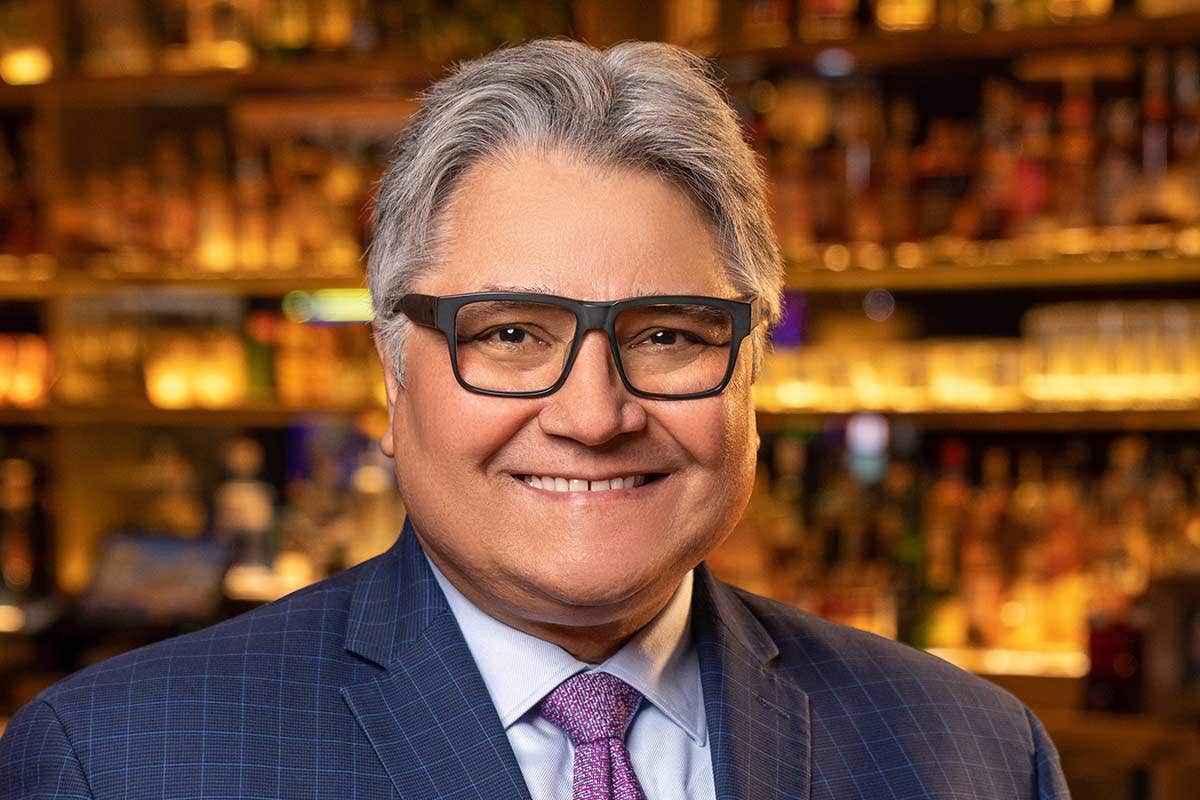 A smiling man with gray hair and glasses, wearing a suit and pink tie, in front of a blurred background of bottles on shelves.