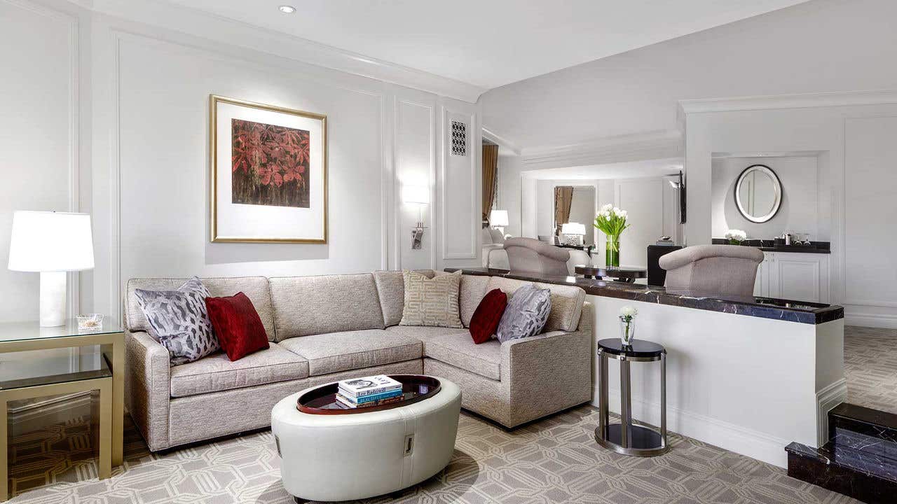 Hotel room with a couch, coffee table, and side table with a lamp. A king bed and two armchairs visible behind the couch on an elevated floor.