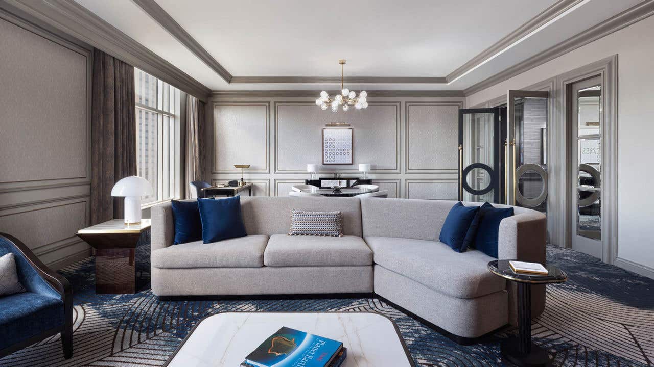 A hotel living room with a couch, dining table, desk, and a set of open double doors on the right.