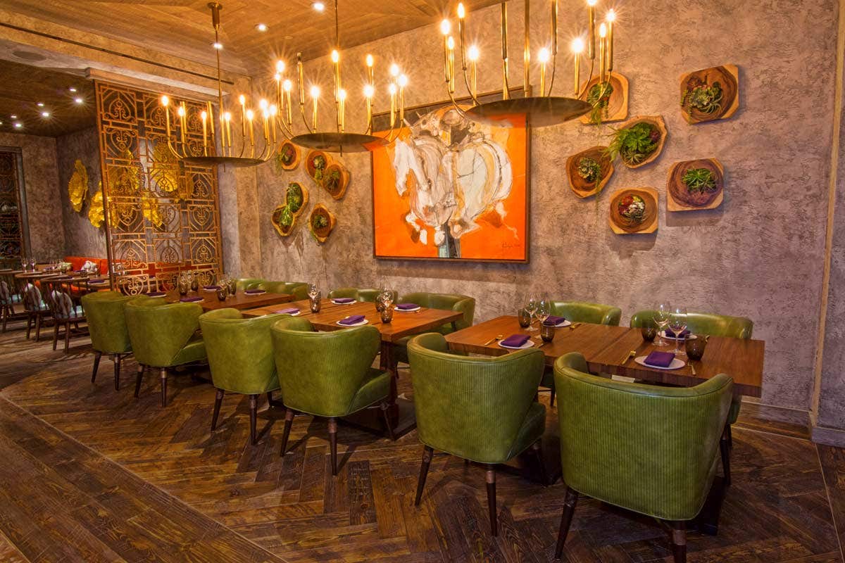 Cozy restaurant with green chairs, wooden tables, hanging planters, chandeliers, and a vibrant horse painting on the wall.