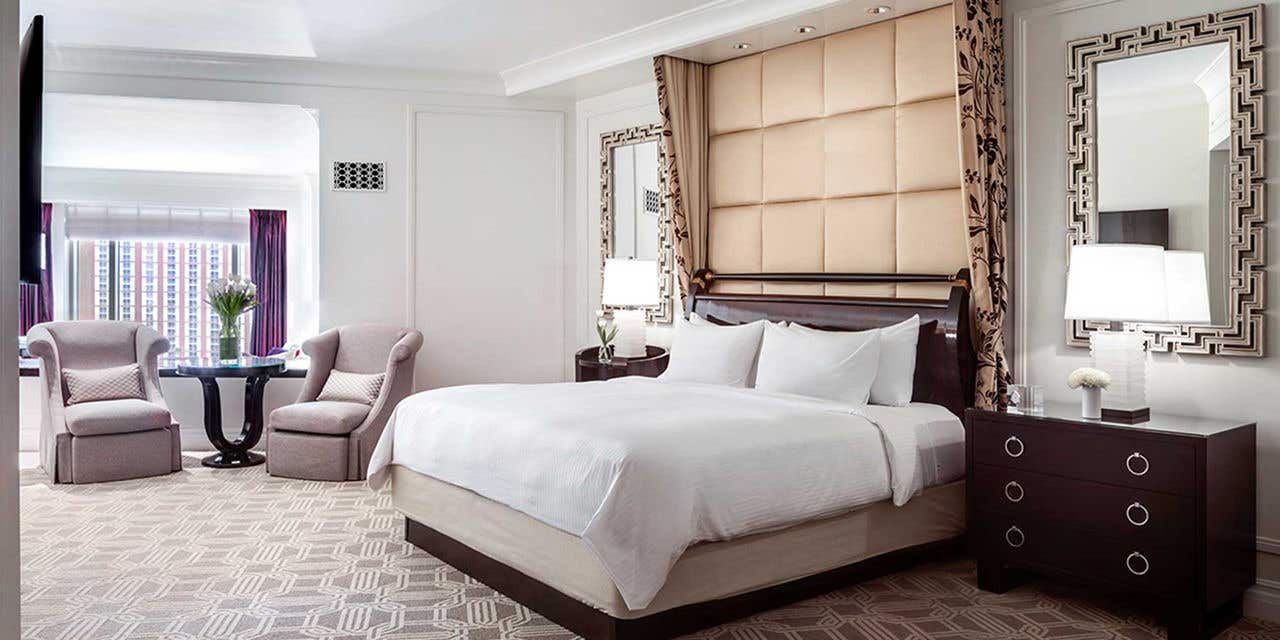Hotel room with a king bed, plush headboard, two armchairs by a round table, and Strip view from the window. Sophisticated decor with mirrored panels and bedside lamps.