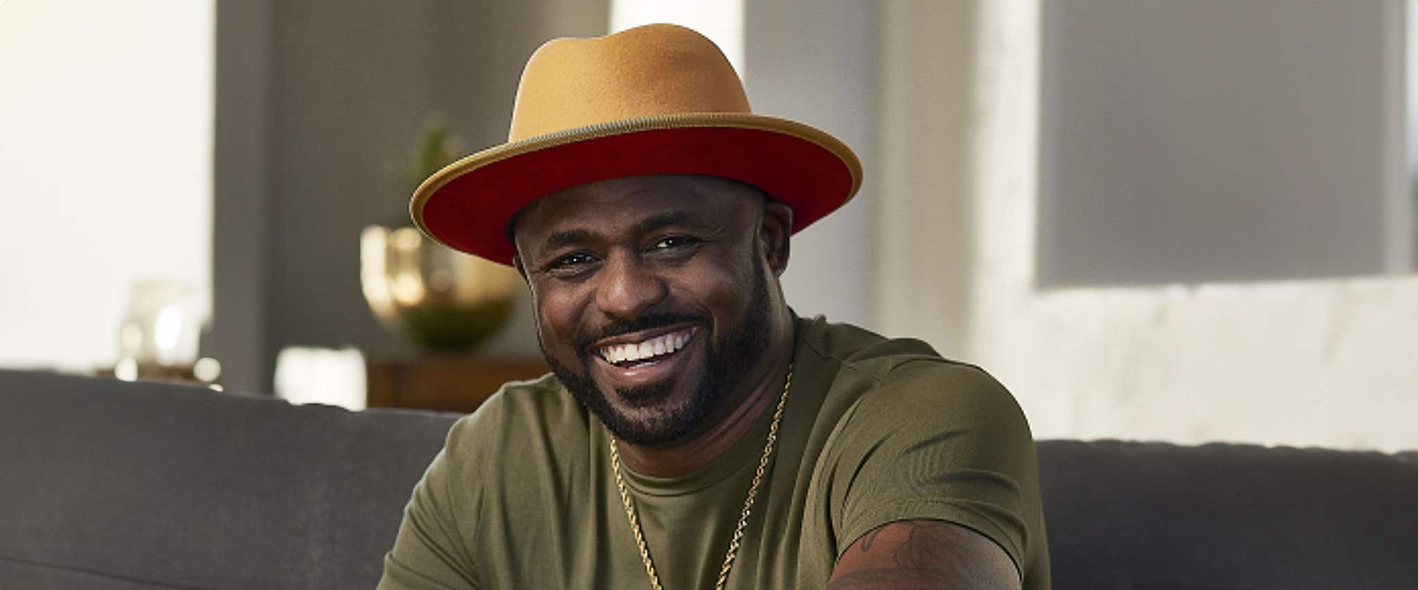 Wayne Brady smiling wearing a tan hat, jeans and green shirt siting on a couch staring straight at the camera. 