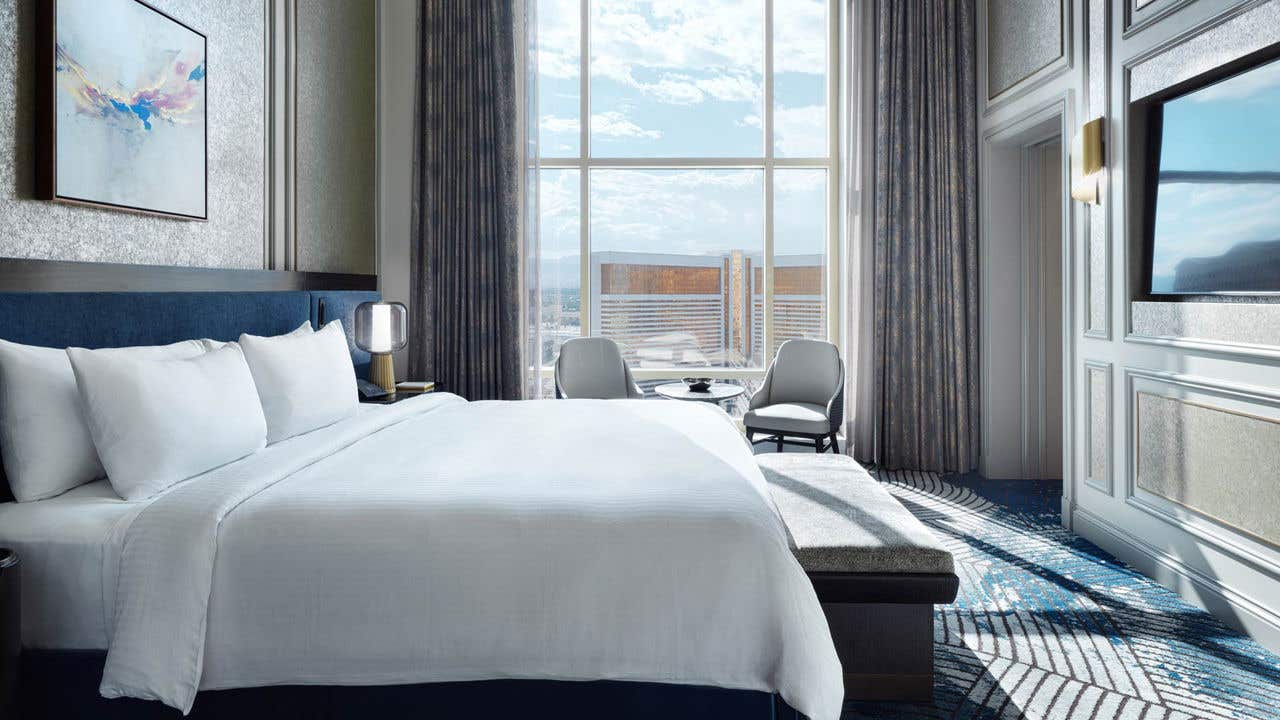 A hotel room with a king bed, bench seat, armchairs, small table, wall-mounted TV, and a tall arched window with a view of the Las Vegas Strip.