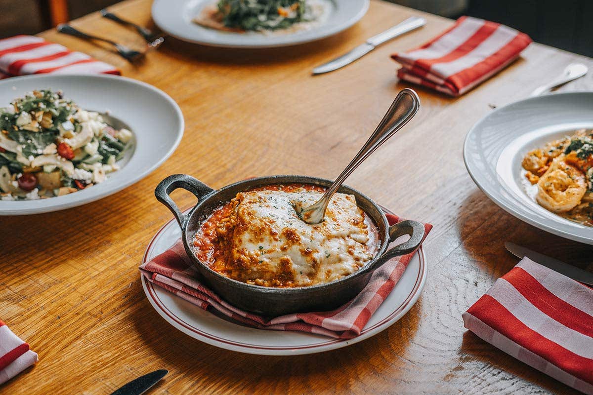 Lasagna in a cast iron skillet with a fork inserted in it, on a table amongst other plates and table settings