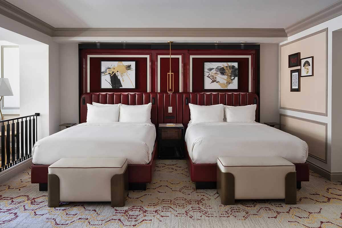 A hotel room with two queen beds, each with a bench at the foot of the bed, a dark red headboard, and modern art on the wall above the beds.